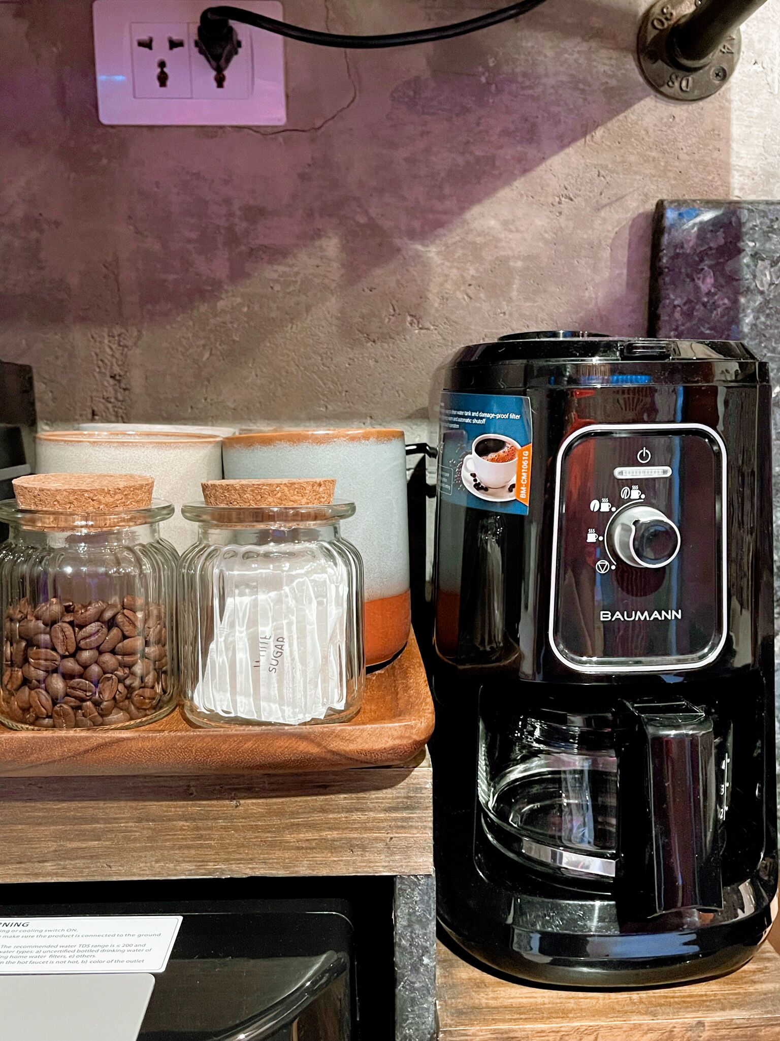 Una cafetera sobre una encimera de madera, con tarros de granos de café y azúcar.