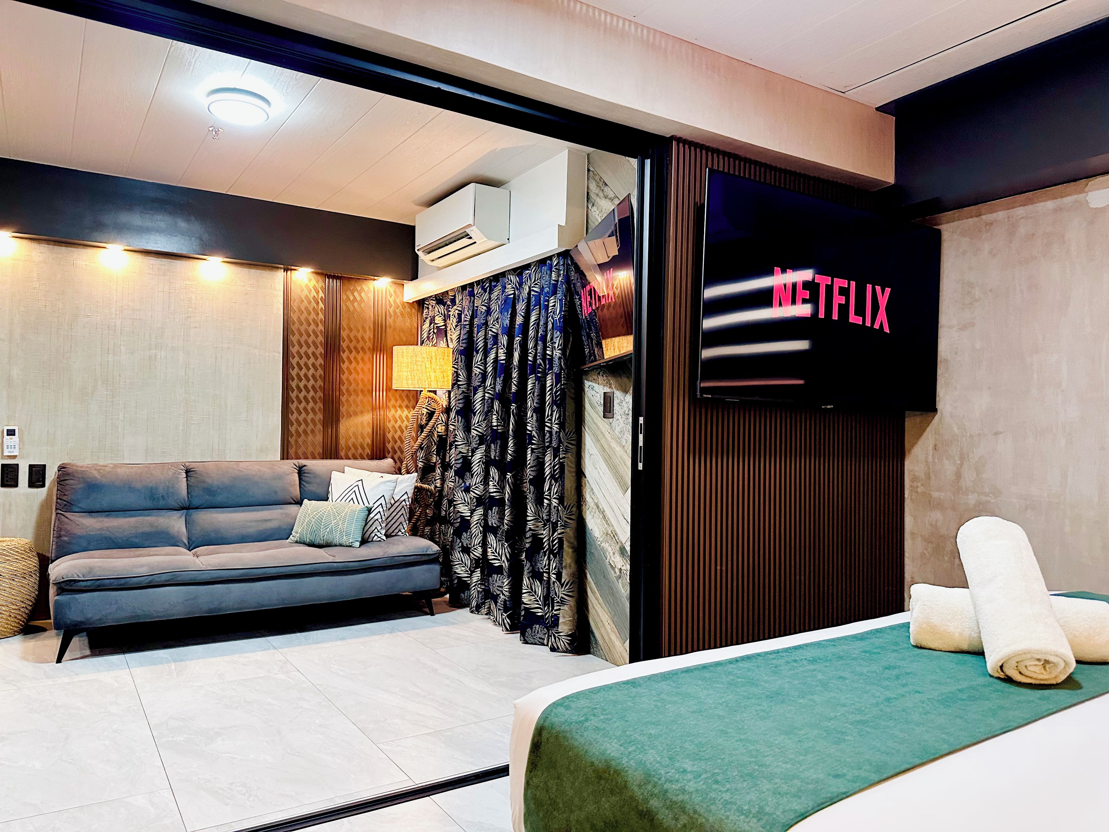 A modern hotel room with a sofa, a bed with a green bedspread, a wall-mounted TV displaying 'NETFLIX,' and a patterned curtain.