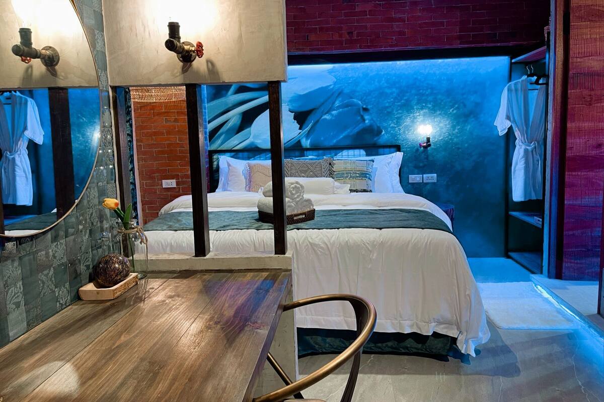 A modern hotel room with a bed, a wooden desk, a mirror, and a brick wall.