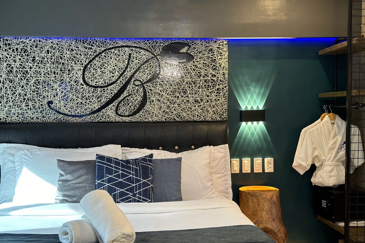 Ein modernes Hotelzimmer mit einem großen Bett, einem Kopfteil mit geometrischem Muster, einem Nachttisch aus Holz und einem Bademantel, der an einem Haken hängt.