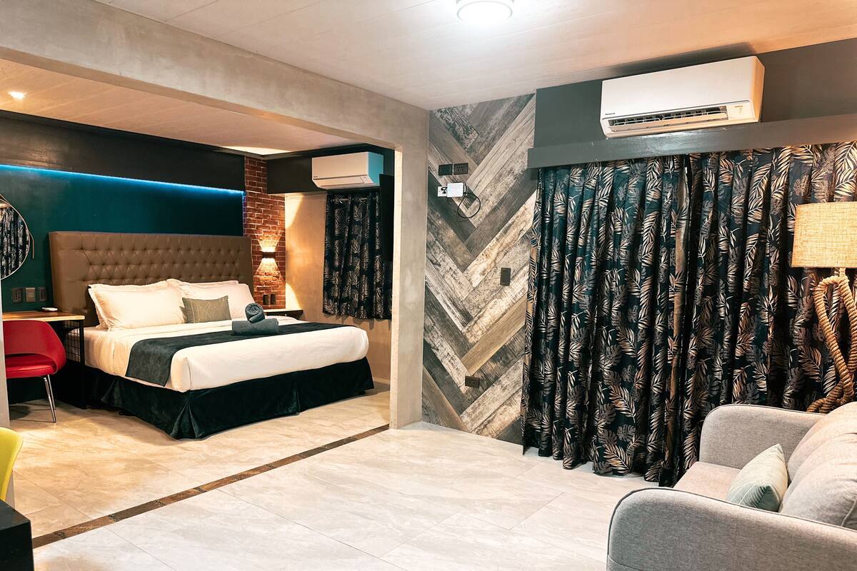 A modern hotel room with a large bed, a desk, a red chair, a sofa, and a patterned curtain.