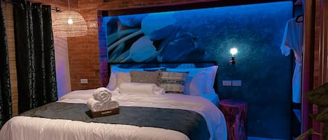 A bedroom with a brick wall, a bed with white bedding, a decorative wall hanging, and a small side table.