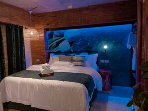 A bedroom with a brick wall, a bed with white bedding, a decorative wall hanging, and a small side table.