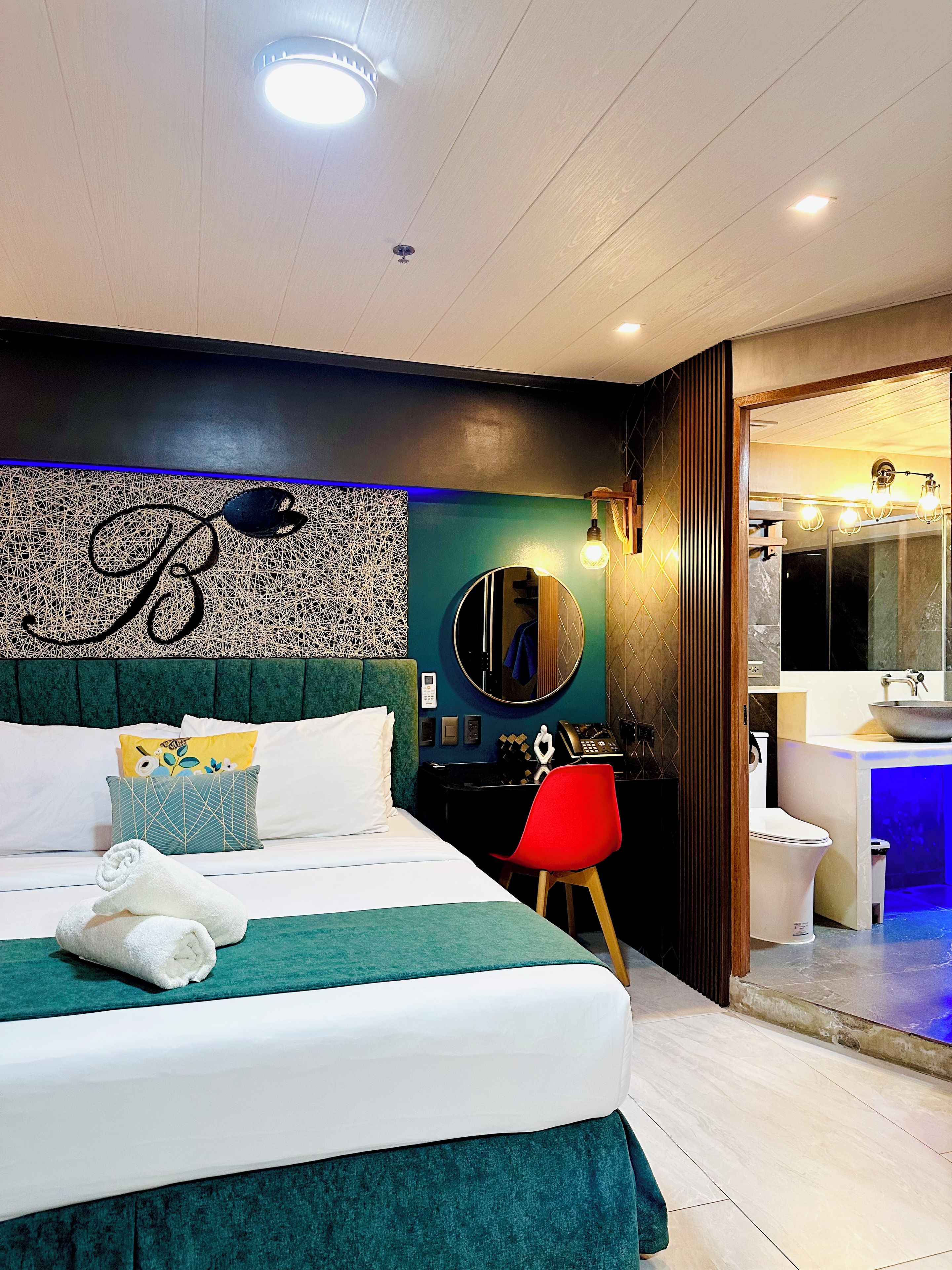 A hotel room with a bed, a desk, a red chair, a bathroom with a sink, and a mirror.