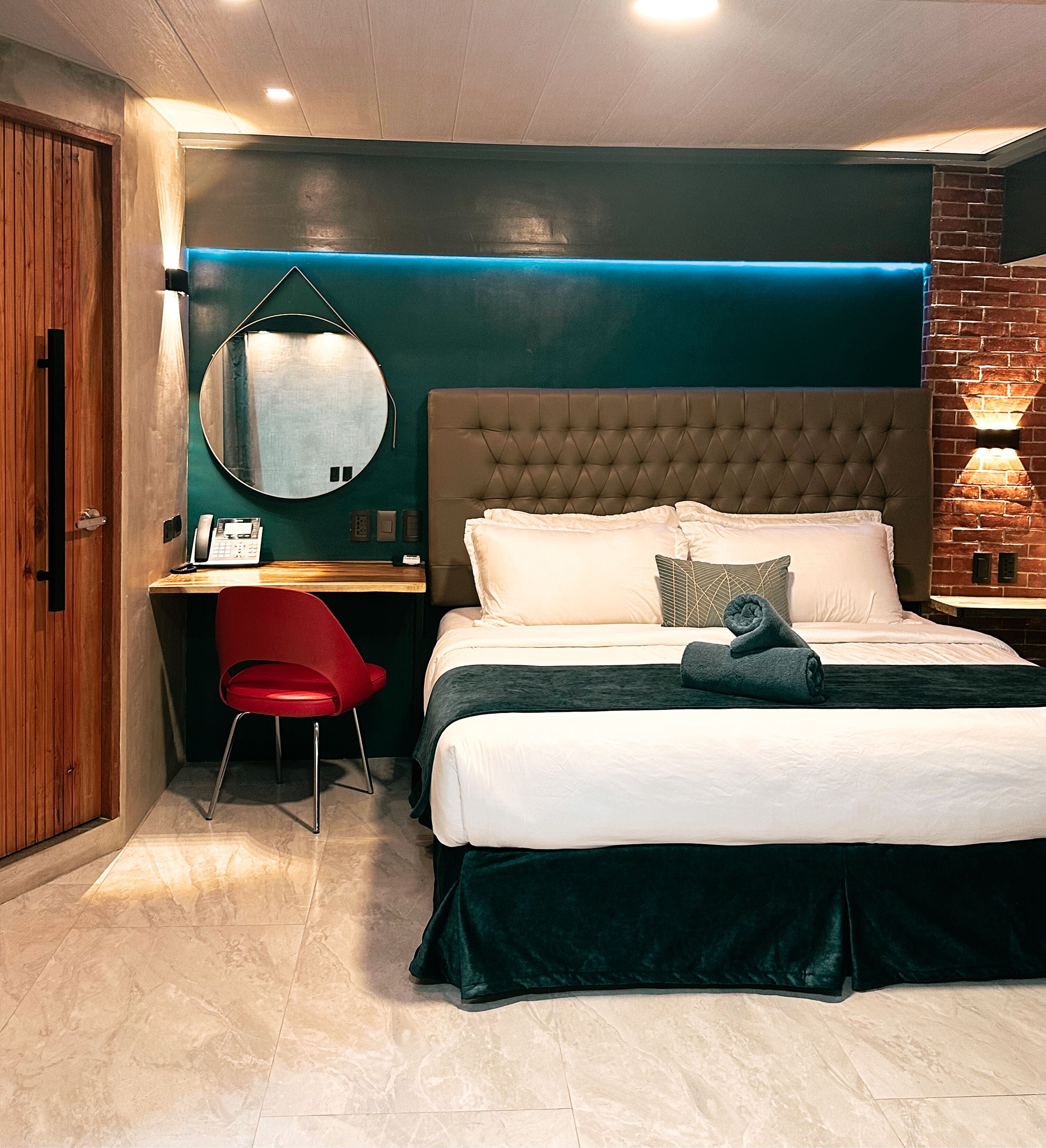 A modern hotel room with a large bed, a desk, a red chair, and a round mirror.