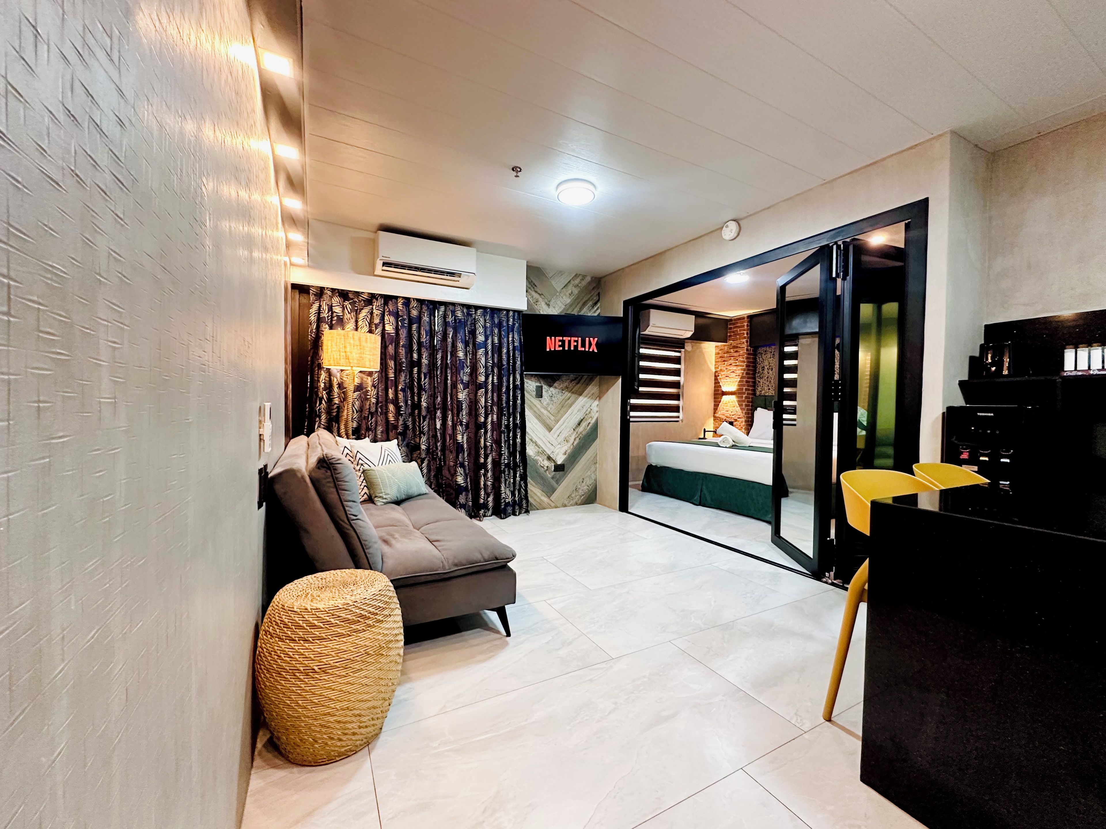A modern hotel room with a sofa, a small round ottoman, a bed with a green bed skirt, a black desk with a chair, and a wall-mounted air conditioning unit.