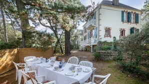 An outdoor dining area with white plastic chairs and a table set for four, surrounded by pine trees and a stone wall.