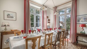 A dining room with a table set for six, wooden chairs, a chandelier, and large windows with red curtains.