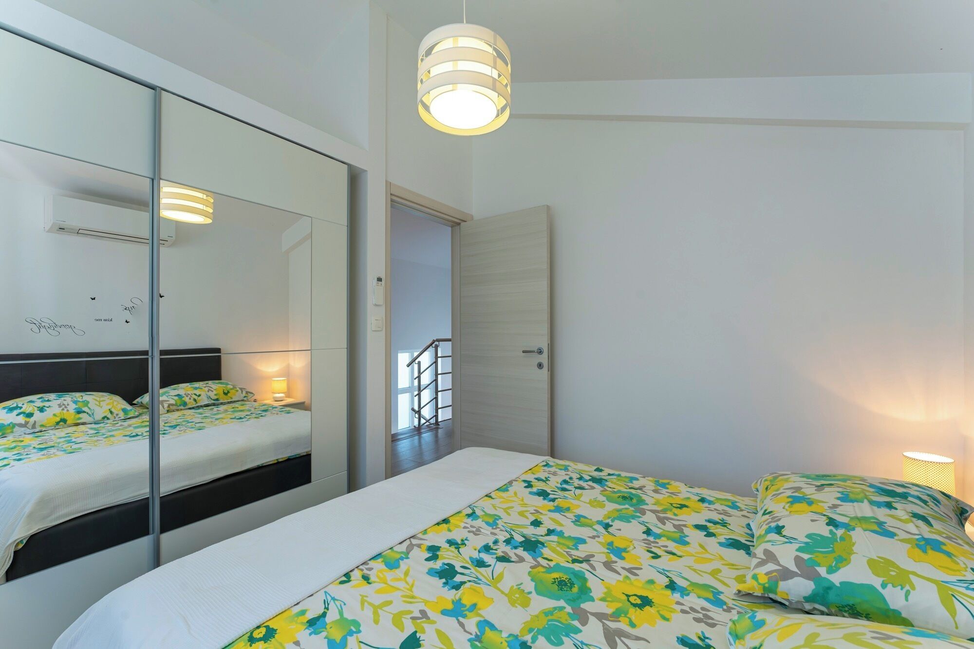 Ein modernes Schlafzimmer mit einem großen Spiegel, einem Bett mit floralem Bettzeug, einem Nachttisch mit Lampe und einer Deckenleuchte.