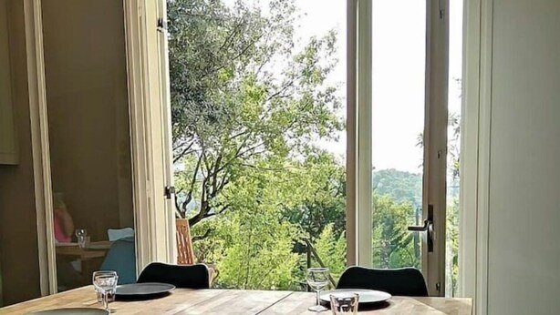 A dining area with a wooden table and four chairs, large windows with a view of greenery, and a door leading outside.