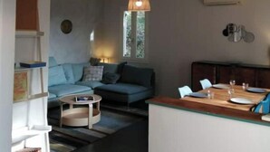 A modern living room with a sofa, a coffee table, and a dining area with a wooden table and chairs. There is a window with a view of greenery outside.