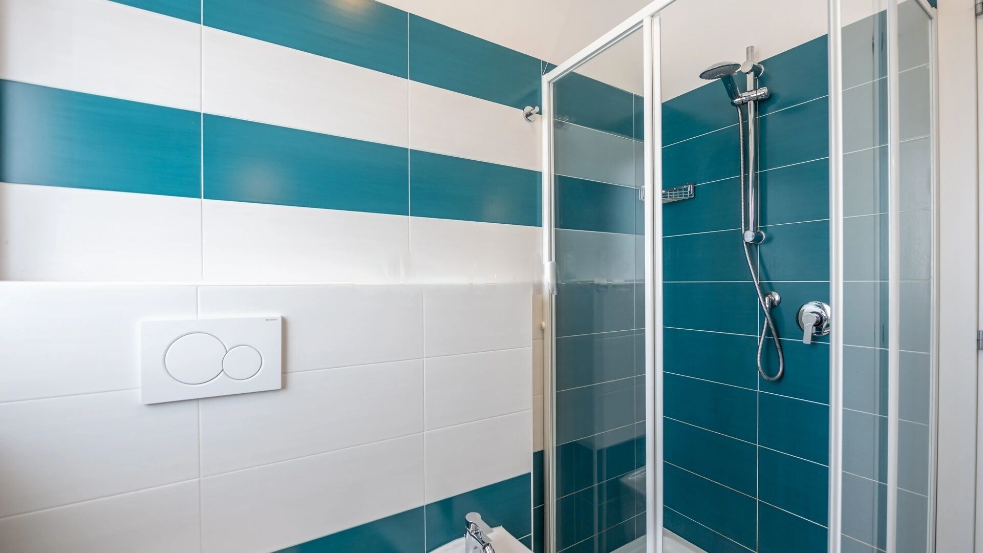 Un baño con cabina de ducha de vidrio, azulejos blancos y turquesa, e interruptor de luz a la vista.