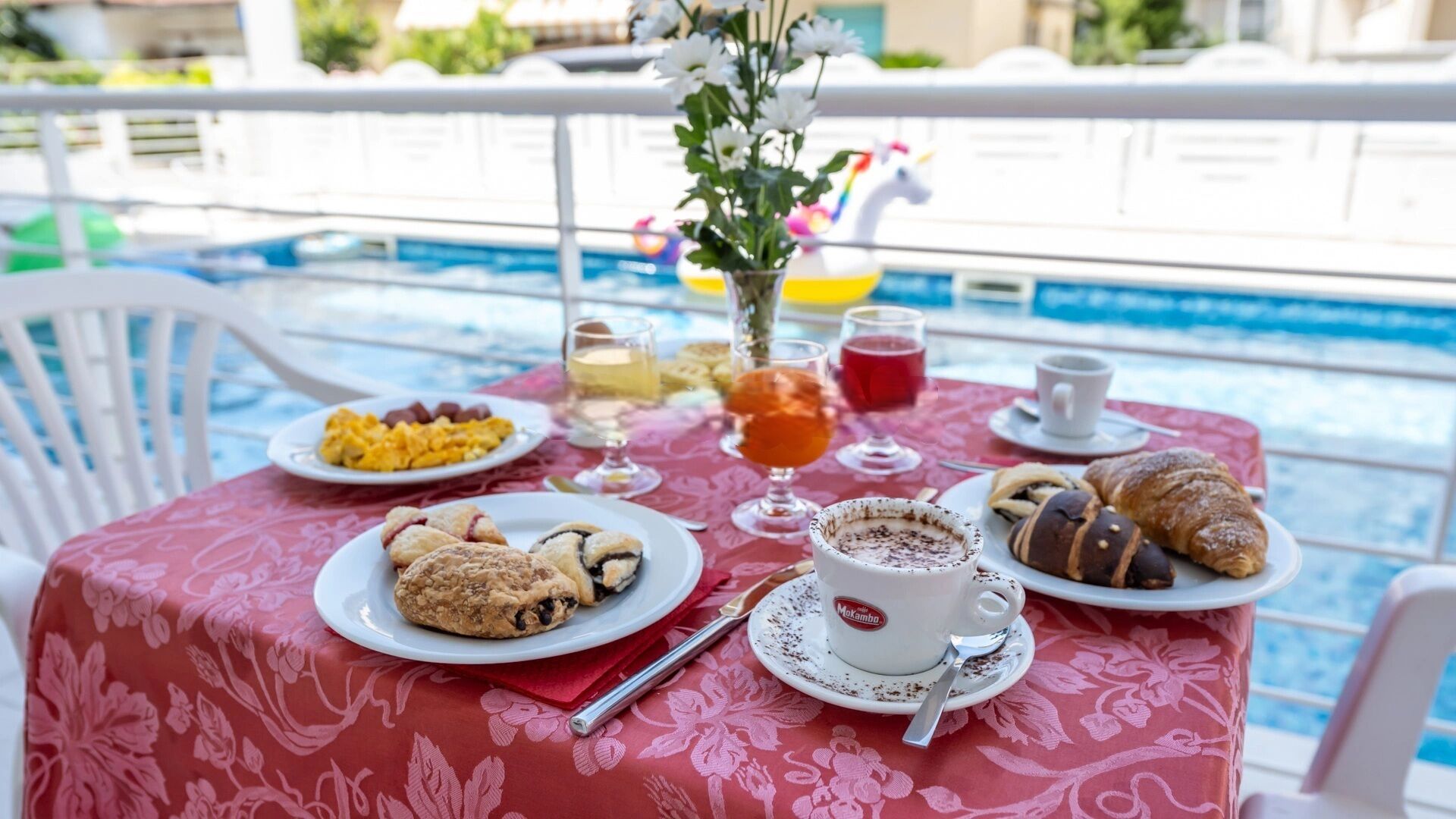 A dining table with breakfast items, including pastries, a cup of coffee, and a glass of juice, by a poolside.