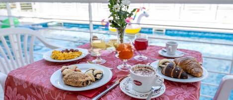 Una mesa de comedor con alimentos para el desayuno, incluyendo bollería, una taza de café y un vaso de jugo, junto a una piscina.