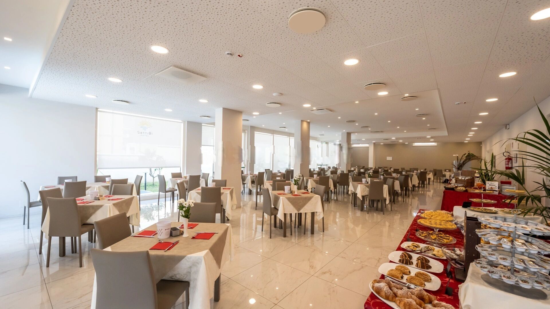 Un comedor amplio con varias mesas y sillas, ventanas grandes y una barra de buffet con diversos alimentos.