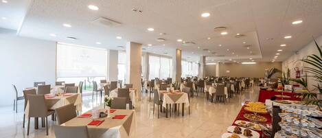 Un comedor amplio con varias mesas y sillas, ventanas grandes y una barra de buffet con diversos alimentos.