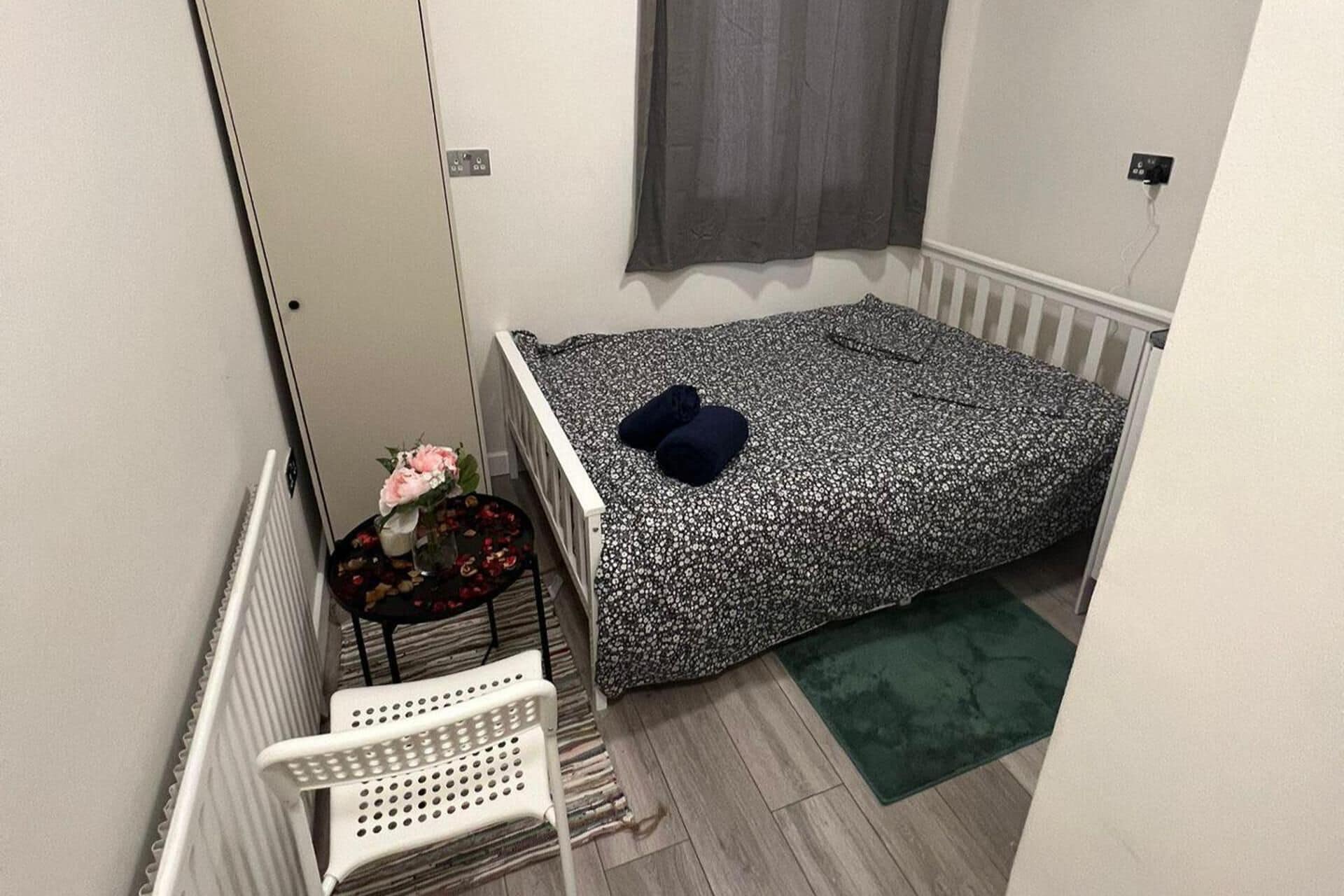 A small, single-bed room with a white radiator, a small table with a vase of flowers, a white chair, and a green rug.
