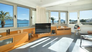 A modern living room with a large flat-screen TV, wooden furniture, and large windows offering a view of the sea and mountains. - Magnific Apt 2 Sea View by JS Villas (Puerto Pollensa)