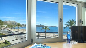 A room with a large window overlooking a beach and the sea, a wooden desk with a magazine, glasses, and two glasses of water. - Magnific Apt 2 Sea View by JS Villas (Puerto Pollensa)