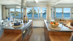 A modern kitchen with a central island, large windows with a view of the sea, and a dining area with white chairs. - Magnific Apt 2 Sea View by JS Villas (Puerto Pollensa)