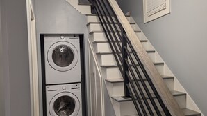 A laundry room with two stacked front-loading washers and dryers, a wooden staircase with black railings, and a geometric light fixture above the staircase.