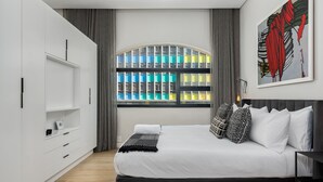 A modern bedroom with a large bed, a wardrobe, a window with a view of colorful buildings, and a painting on the wall. - Luxurious Studio Apartment | Netflix | Wifi (Cape Town)