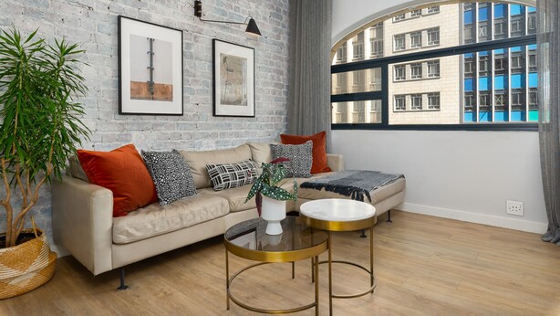 A modern living room with a brick wall, a large window with a view of buildings, a sofa with cushions, a coffee table, and a potted plant. - Luxurious Studio Apartment | Netflix | Wifi (Cape Town)