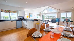 A modern kitchen with white cabinets, a dining area with a wooden table set for six, and a view of the sea through large windows.