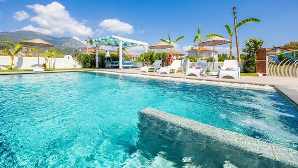 Una piscina cristallina con lettini e ombrelloni, circondata da palme e case.