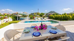 Area pranzo a bordo piscina con tavolo apparecchiato per due, piscina e un pergolato con ombrelloni.