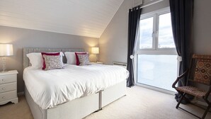 Ein Schlafzimmer mit einem großen Bett, zwei Nachttischen mit Lampen, einem Sessel und einem Fenster mit Vorhängen.