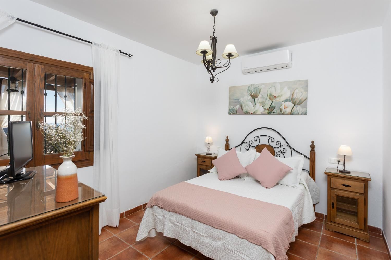 Une chambre à coucher avec un lit, des tables de chevet, une télévision, un lustre et une fenêtre avec des rideaux.