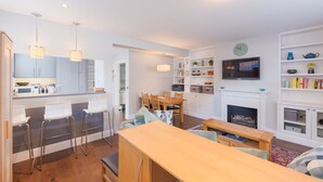A modern kitchen with white cabinets, a dining area with a wooden table and chairs, a TV mounted above a fireplace, and a bookshelf with various items. - Fabulous Fulham - Two Bedroom Apartment, Sleeps 4 (London)