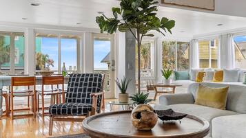A spacious living room with a large window, a wooden coffee table, a patterned armchair, and a grey sofa with yellow cushions.