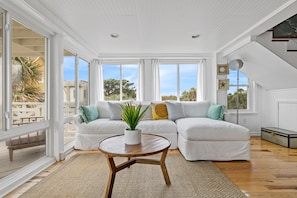 A spacious living room with a white sofa, a round wooden coffee table, and a large window offering a view of the outdoors.