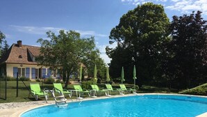 Una piscina con sillas de descanso verdes y sombrillas, rodeada de baldosas de piedra y una casa al fondo.