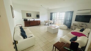 Un saló modern amb sofàs blancs, una taula de centre i una televisió. Hi ha una zona de menjador amb una taula i cadires, i una finestra gran amb cortines.