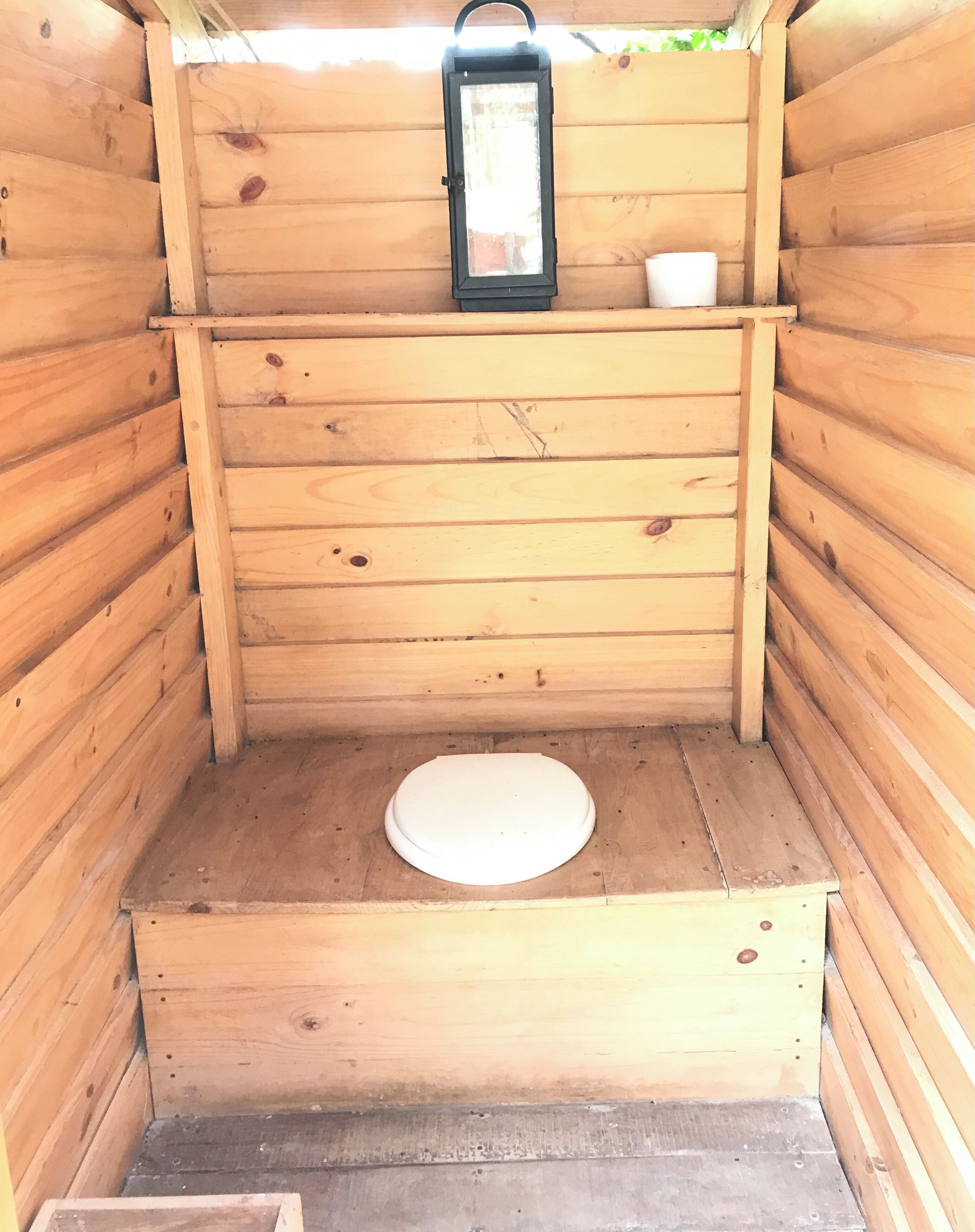 A wooden outhouse with a white toilet, a lantern, and a small shelf with a cup.