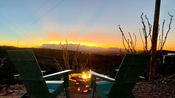 Twee groene stoelen gericht op een kampvuur, met een zonsondergang en bergen op de achtergrond.