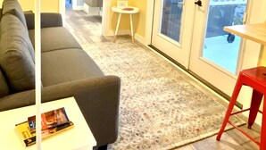 Een woonkamer met een bank, een kleine tafel, een rode kruk en uitzicht door een glazen deur.