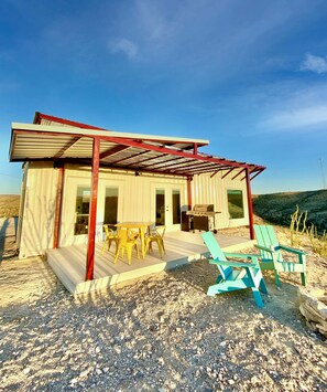 A small, single-story building with a covered patio, outdoor seating, and a barbecue grill.