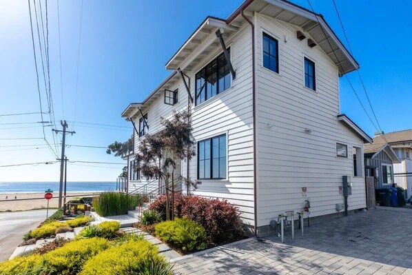 A two-story white house with a covered porch, a small garden, and a view of the beach.