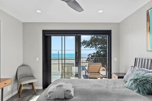 A bedroom with a bed, a chair, a small table, and a view of the beach through a sliding door. - Luxury Twin Lakes Beachhouse (Santa Cruz)