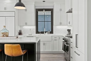 Une cuisine moderne avec des meubles blancs, un luminaire suspendu noir, une grande fenêtre et un plancher en bois.