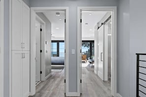 A modern hallway with white doors, a bed in the background, and a view of the outdoors.