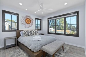 A modern bedroom with a large bed, a bench, and three windows offering a view of the outside. - Luxury Twin Lakes Beachhouse (Santa Cruz)