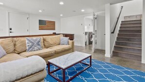 Una sala moderna con un sofá color beige, una mesa de centro de mármol y un tapete azul estampado. Una escalera de madera con barandilla de metal conduce a un piso superior.
