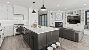 Una cocina moderna con armarios blancos, una isla central con taburetes, horno empotrado y un televisor de pantalla plana grande sobre la isla.