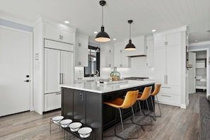 A modern kitchen with white cabinetry, a central island with bar stools, pendant lighting, and a visible refrigerator.