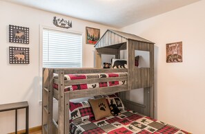 Un lit superposé avec une structure extérieure en bois, une fenêtre avec des stores et des décorations murales comprenant des motifs d’animaux et un coussin à l’effigie d’un ours.
